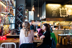 People enjoy drinks at the bar, while an open kitchen is featured in the background at Cafe Bar in West Washington Park. 